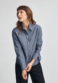Camicia a maniche lunghe con bottoni in tessuto blu scuro. Presenta un colletto classico, un logo sul petto, bottoni bianchi e una forma leggermente aderente.