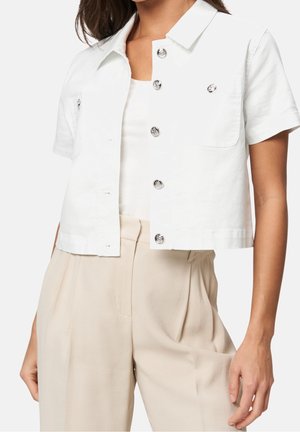 Kvinde iført en hvid kortærmet, knaplukket cropped jakke over en hvid tanktop, kombineret med højtaljede beige bukser med plisseringer.