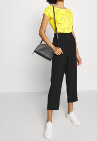 Person klädd i en gul mönstrad topp, svarta korta vida byxor, vita sneakers och bärande på en svart läder axelväska.