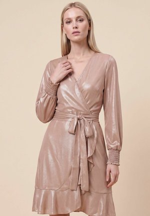 Kvinde iført en skinnende lyserød slå-om-kjole med lange rynkede ærmer og en bundet bue i taljen, stående mod en neutral beige baggrund.
