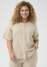 Beige overhemd met korte mouwen en knoopsluiting, gemaakt van een gestructureerde stof en voorzien van een borstzak. Het heeft een ronde halslijn en zichtbare knopen.