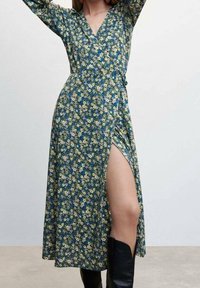 Kleid mit floralem Druck in Grün mit blauen und gelben Blumen. Verfügt über einen tiefen V-Ausschnitt, lange Ärmel und einen Seitenschlitz. Wird mit schwarzen Stiefeln getragen.