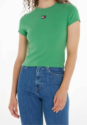 Vrouw draagt een groen kort mouw crop top met Tommy Jeans-logo en een high-waisted blauwe spijkerbroek tegen een witte achtergrond.