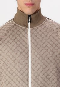 Brah taktilna zip up jakna z visokim ovratnikom in belim zadrgo. Tkanina ima geometrijski vzorec in strukturirano površino.