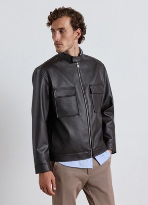 Mann trägt eine schwarze Lederjacke mit vorderen Klapptaschen, zugezippt, über einem blauen Hemd und beigen Hosen, schaut zur Seite.