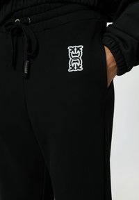 Zwarte joggers met een trekkoord in de taille, voorzien van een zijzak en een wit geborduurd logo op de dij. Glad stofoppervlak.