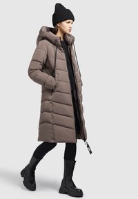 Lange gewatteerde jas in taupe met een capuchon, voorzien van verticale stiksels, ritsen en koorden. Gekocht met zwarte legging en combat boots.