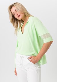 Hellgrünes, kurzärmliges Oberteil aus weichem Stoff mit V-Ausschnitt und transparenten Akzenten an den Ärmeln. Getragen mit weißen Hosen, die seitliche Taschen haben.