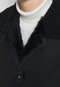 Manteau noir avec un col en fausse fourrure pelucheux, fermeture à boutons et texture lisse. Superposé sur un col roulé blanc.