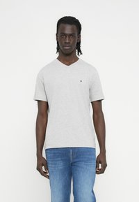 Γκρι βαμβακερό t-shirt με V-λαιμόκοψη και κοντά μανίκια. Διαθέτει μικρό λογότυπο σε κόκκινο, άσπρο και μπλε στο στήθος. Φορεμένο με μπλε τζιν.