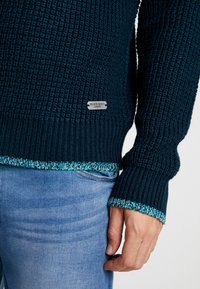 Mörk teal stickad tröja med ribbad textur, med kontrasterande mönstrad fåll och ärmkanter. Silvermärke med "Brave Soul"-logotyp synlig.