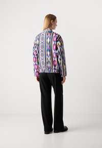 Vrouw die wegdraait en een levendige, kleurrijk patroonjacket draagt met zwarte, wijde broek en zwarte schoenen, op een effen lichte achtergrond.