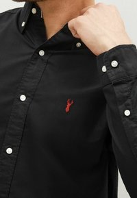 Zwart overhemd met knopen en een kraag, met witte knopen en een klein rood kreeftborduursel op de linkerborst. Gemaakt van katoen.