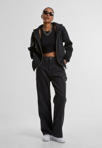 Felpa nera con zip sopra un crop top nero, abbinata a pantaloni larghi neri con tasche. Gli accessori includono occhiali da sole e una collana a catena.