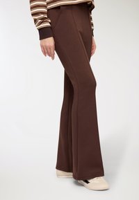 Pantaloni marroni a zampa in tessuto morbido con tasche laterali e dettagli di cuciture visibili, abbinati a scarpe da ginnastica di colore chiaro.