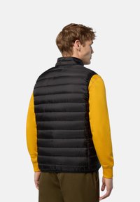 Gilet en duvet noir avec un rembourrage horizontal, un col montant et une texture lisse, porté par-dessus une chemise à manches longues jaune.