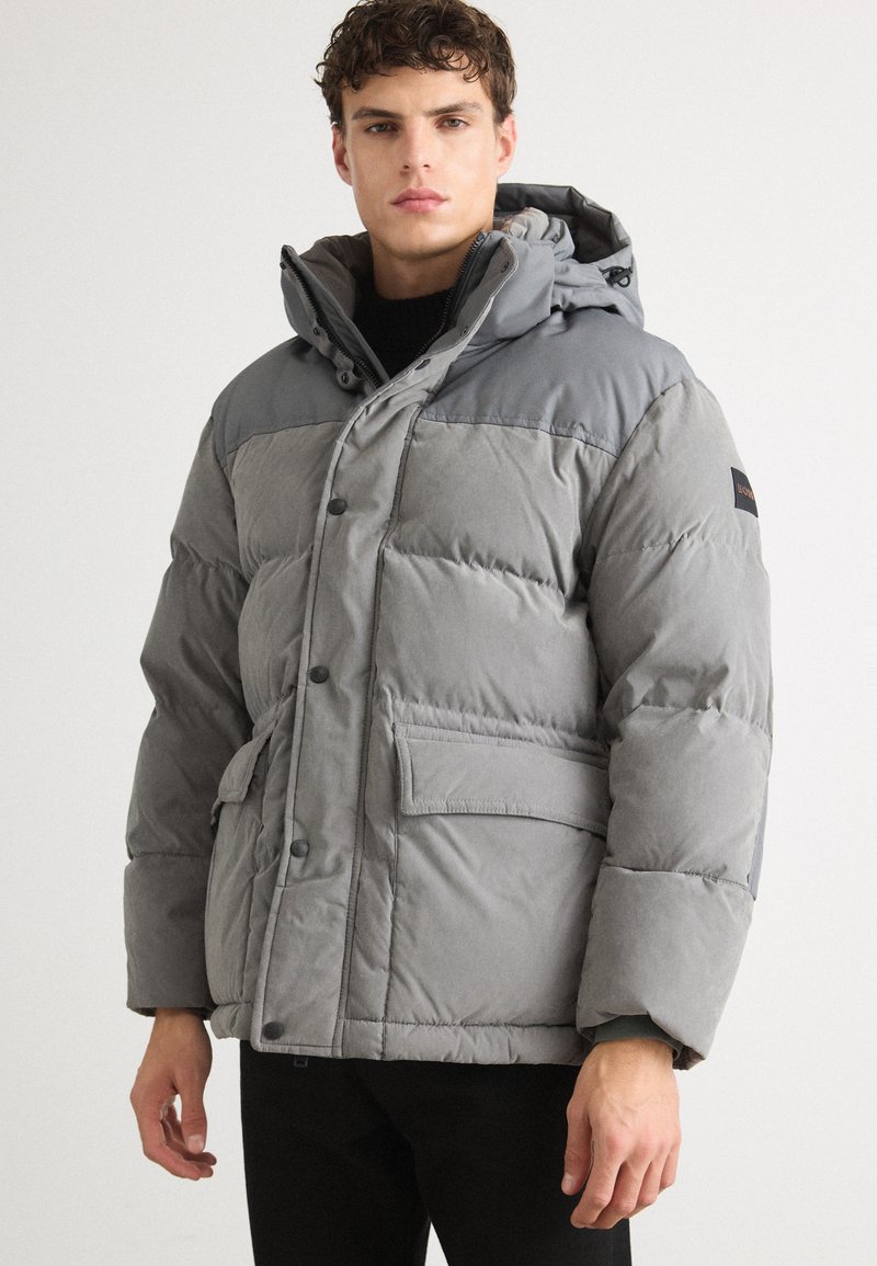 Chaqueta acolchada gris con capucha, cierre frontal de botones y un gran bolsillo de parche. Textura suave con un diseño de hombros en contraste.