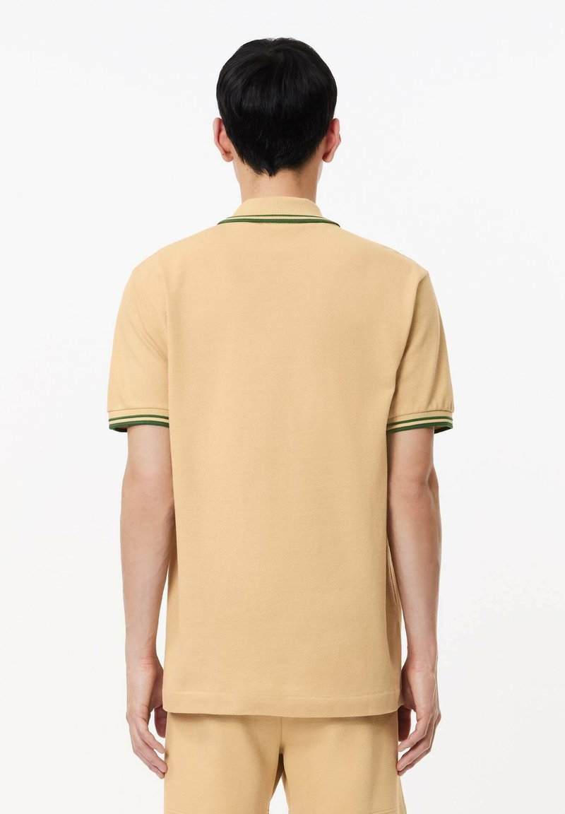 Camisa polo beige con una textura lisa, de manga corta y con un cuello acanalado verde. La parte trasera muestra un corte recto y un diseño minimalista.