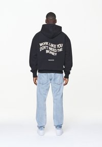 Hombre con sudadera negra con el texto "TRABAJA COMO SI NO NECESITARAS EL DINERO" y jeans de mezclilla azul claro, de pie de espaldas sobre un fondo blanco.