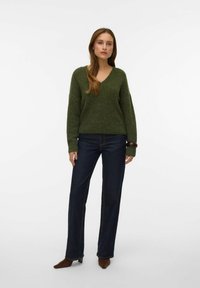 Grüner, strukturierter Strickpullover mit V-Ausschnitt und langen Ärmeln, kombiniert mit dunklen, geraden Jeans und braunen Schuhen mit Absatz.