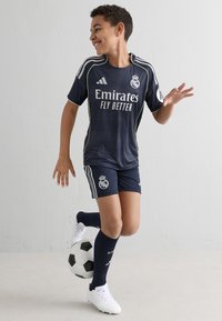 Equipamento de futebol do Real Madrid em azul marinho com detalhes em branco, apresentando o logo "Emirates Fly Better" e meias a condizer, acompanhado de uma bola de futebol em preto e branco.