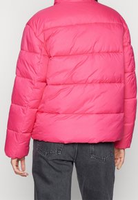 Henkilö, joka pukee yllään kirkkaan vaaleanpunaisen pehmustetun takin ja tummanharmaat farkut, kuvattuna takaa päin, yhdellä kädellä näkyvissä ja yhdellä sormella on side.