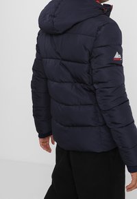 Marinblå puffjacka med huva, med kviltade sektioner och en logotypapplikation på vänster ärm. Tyget ser slätt och texturerat ut.