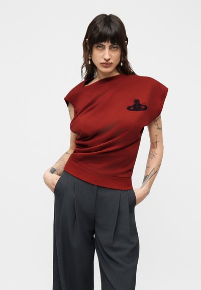 Νεαρή γυναίκα με σκούρα μαλλιά, ντυμένη με κόκκινο αμάνικο τοπ με λογότυπο και σκούρο παντελόνι με φαρδιά μπατζάκια, με τα χέρια στις τσέπες, στέκεται μπροστά σε απλό φόντο.