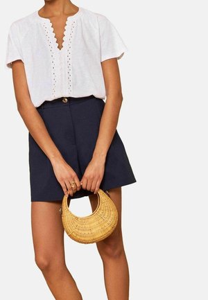 Femme portant un chemisier blanc à col en V festonné et un short taille haute bleu marine, tenant un sac à main rond tissé avec des anses courtes.