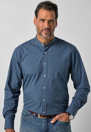 Mann mit kurzen dunklen Haaren und Bart, trägt ein blau gemustertes Hemd mit Mandarinkragen und blaue Jeans, Hand in der Tasche.