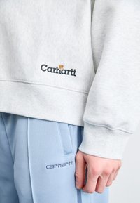 Sweatshirt gris clair avec des cuffs côtelés, arborant le logo "Carhartt" brodé. Pantalon bleu avec poches latérales et broderie "Carhartt" assortie.