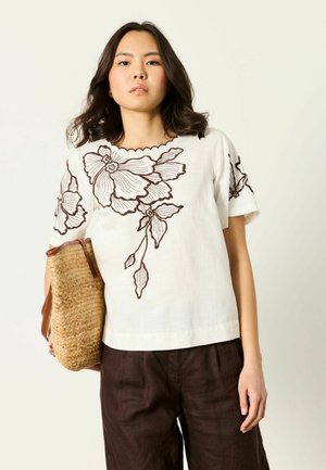 Kobieta ubrana w białą bluzkę z dużą brązową kwiatową haftem, ciemnobrązowe spodnie i trzymająca plecioną torbę z trawy.