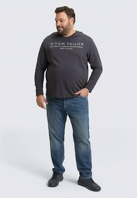 Dark gray long-sleeve shirt with white text, paired with blue jeans and black shoes. Model stands facing slightly left.