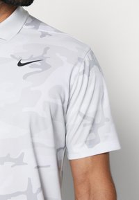 Muž v světlém šedém maskáčovém polosvětlém tričku s krátkým rukávem a černým logem Nike swoosh na prsou.