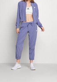 Ensemble de sport violet clair comprenant une veste courte zippée et un pantalon de jogging assorti. Pull et pantalon en tissu lisse avec poignets élastiques.