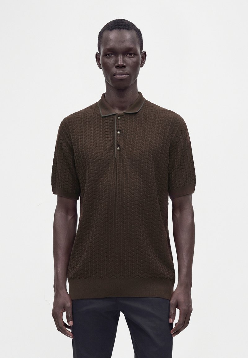 Homme à la peau foncée portant un polo marron texturé à manches courtes et un pantalon foncé, se tenant devant un fond clair uni.
