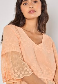 Pfirsichfarbene bestickte Bluse mit V-Ausschnitt, gewellten Kanten und transparenten, ausgestellten Ärmeln, die mit floralen Mustern und strukturierter Stoffqualität versehen sind.
