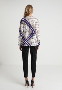 Blusa a maniche lunghe con stampa floreale su sfondo bianco e accenti diagonali viola; abbinata a pantaloni neri aderenti e tacchi a punta.
