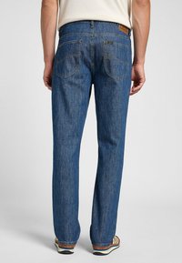 Jeans de mezclilla azul con un ajuste recto, que cuentan con un parche de cuero en la parte posterior, diseño de cinco bolsillos y detalles de costura visibles.