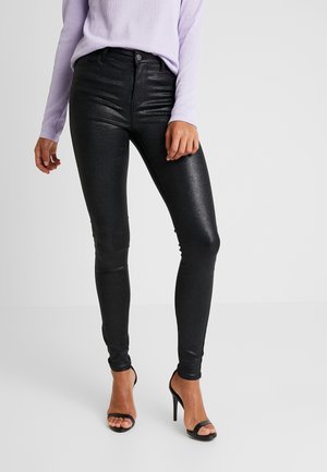 Kobieta nosząca błyszczące czarne dopasowane spodnie, liliową bluzkę z długim rękawem oraz czarne sandały na wysokim obcasie, stojąca na jednolitym tle.