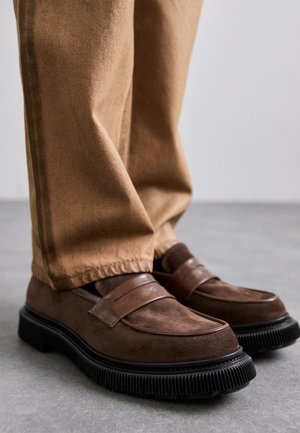 Brown suede loafers with a thick black rubber sole and decorative strap detail. Worn with beige trousers.