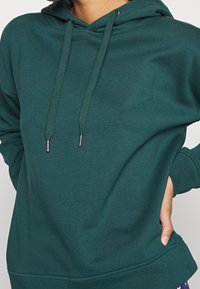 Grön hoodie med dragsko på huvan, har en mjuk textur, känguruficka och ribbade ärmslut. Materialet verkar slätt och hållbart.