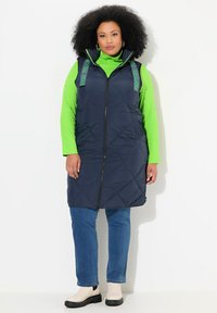 Granatowa puchowa kamizelka z zielonymi paskami z napisem "DOBRZE CZUJ SIĘ DOBRZE", noszona na jasnoprzewym golfie i niebieskich dżinsach. Gładka faktura, do kolan.