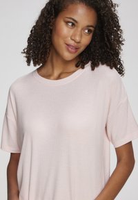 Kortsade lokkidega naine, kellel on seljas helepunane soonikust lühikese varrukaga pluus, vaatab veidi küljele ja naeratab õrnalt.