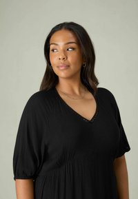 Svart, texturerad klänning med V-ringning och korta ärmar; har delikata utskärningar längs halsen.