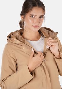 Giacca leggera beige con cappuccio, chiusura con zip e bottoni a pressione. Il materiale sembra liscio; presenta un rivestimento interno trapuntato.