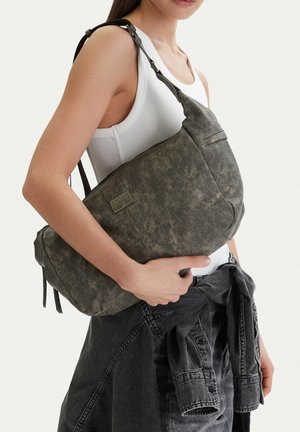 Žena v bílém tílku nese velkou tmavě šedou tašku přes rameno a má kolem pasu uvázanou černou džínovou bundu.