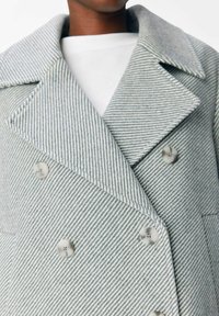Manteau rayé gris et blanc avec de grands revers et une fermeture à double boutonnage. Présente des boutons ronds et des détails de couture visibles.