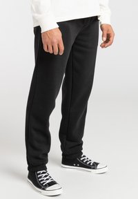 Black sweatpants made of soft fabric, featuring a tapered leg design, elastic cuffs, and side pockets. Paired with black high-top sneakers.