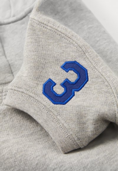 Vaaleanharmaa kangas, johon on kirjailtu sinisellä langalla numero "3" taitetun vaatteen hihassa.